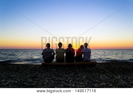 A group of people sitting on the beach and watching the sunset. Silhouettes of people against the sun. Clear blue sky. Stone beach. Holidays by the sea. Georgia