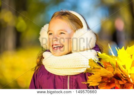 Happy little girl in earflaps with autumn leaves in the park