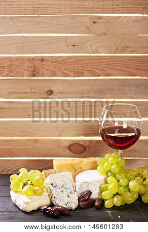 The Glass of red wine served with grapes date fruits and the variety of cheese on a black wooden table with the wooden board on a background with a ray of light shining through it. Empty space