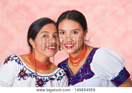 Beautiful hispanic mother and daughter wearing traditional andean clothing, embracing while posing happily together interacting for camera, pink studio background.