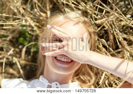 nature, summer holidays, vacation and people concept - close up of happy young woman lying on cereal field and covering face by hand