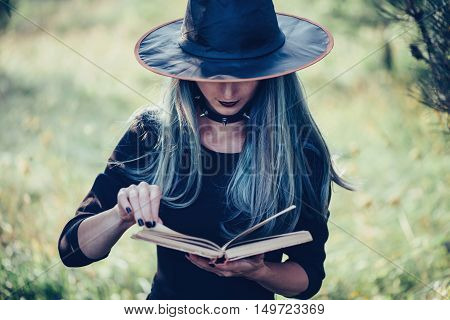 Young woman in costume of witch reading a book of magic outdoor close-up front view. Theme of Halloween and magic