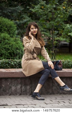 Young beautiful brunette woman in beige coat sitting on a bench in the autumn park