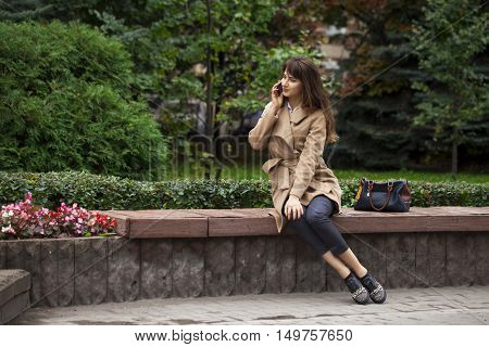 Young beautiful brunette woman in beige coat sitting on a bench in the autumn park