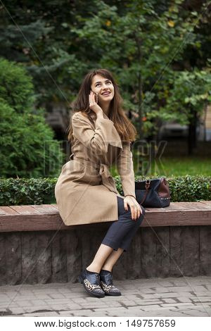 Young beautiful brunette woman in beige coat sitting on a bench in the autumn park