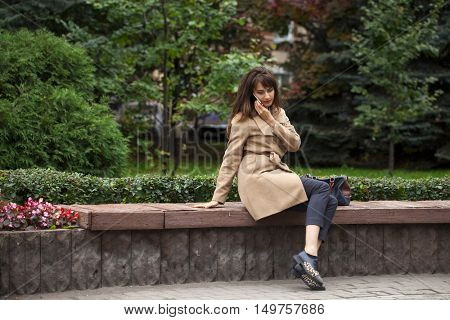 Young beautiful brunette woman in beige coat sitting on a bench in the autumn park