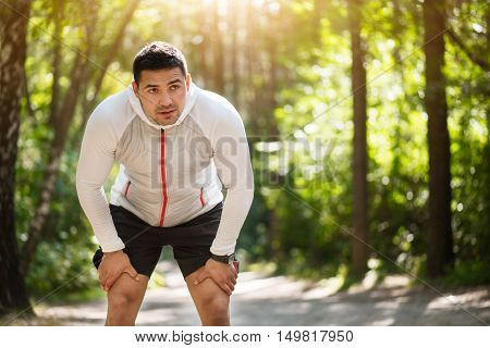 Long distance running. Pleasant tired young man bending forwards and touching his legs while running in the park
