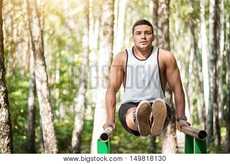 Workout in the park. Handsome athletic serious man leaning on parallel bars and stretching his legs forward while holding his body in the air