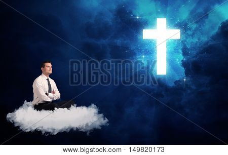 Caucasian businessman sitting on a white fluffy cloud looking and wondering at a big bright glowing cross