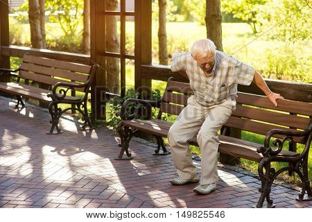 Senior man holding his back. Old male near park bench. Strong pains in spine. Need of medication and therapy.
