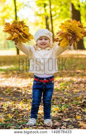 Joyful little girl in park in autumn with leaves in her hands in white knitted cap sunny day