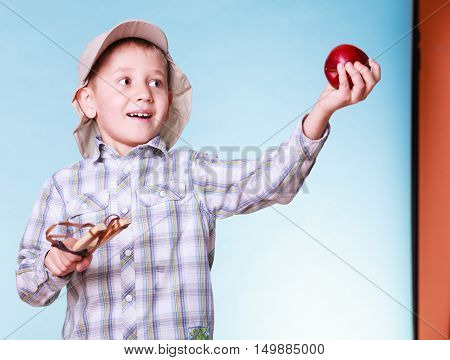 Nature and free time prankster little boy. Child have fun with wooden sling shot and fruit hold apple.