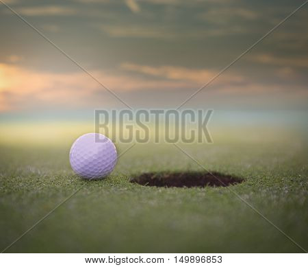 Golf ball sitting on a golf course putting green next to the hole. with a beautiful sunset.