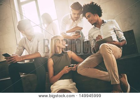 Students life. Low angle view of cheerful young people communicting while holding different gadgets and sitting close to each other on steps