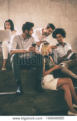 Modern students. Group of cheerful young people communicting while holding different gadgets and sitting close to each other on steps
