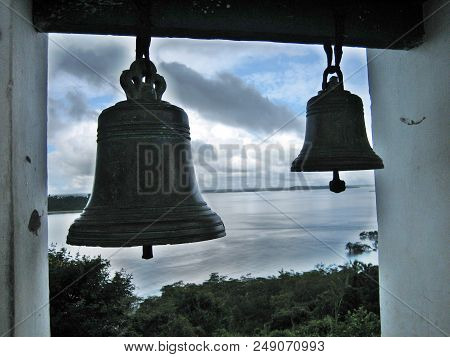 Two bells are hanging over an ocean bay in front of a cloudy sky.