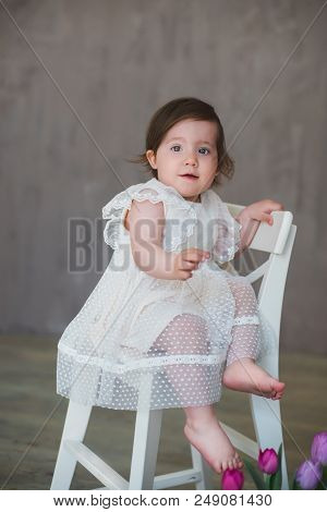 Beautiful Smiling Little Girl In White Lace Dress Among Tulips, Sitting On A White Chair.