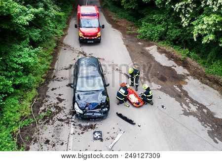 Three Unrecognizable Firefighters Helping A Young Injured Woman Lying On The Road After An Accident.