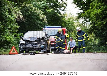 Three Unrecognizable Firefighters Helping A Young Injured Woman Lying On The Road After An Accident.