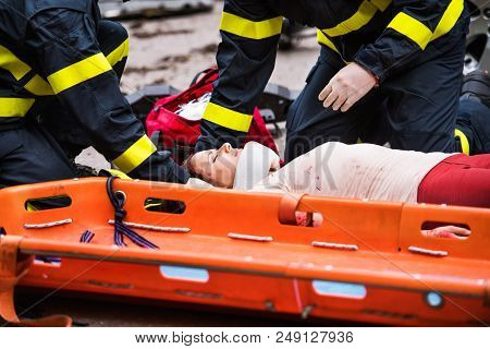 Unrecognizable Firefighters Helping A Young Injured Woman Lying On The Road After An Accident. A Pla