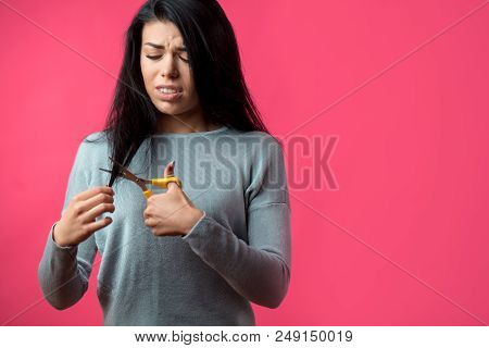 Upset Beautiful Girl Treaming Her Long Black Hair With Yellow Scissors Isolated On The Pink Backgrou