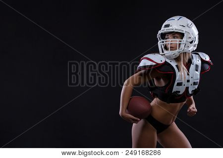 Sportive Serious Woman In Helmet Of Rugby Player Holding Ball In Stuio On Dark Background. Girl Spor