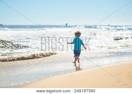 Happy Boy In Blue Swimsuit Running On The Beach In The Waves, Family On Vacation, View From Behind