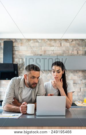 Portrait Of Shocked Married Couple Looking At Laptop Screen Together At Counter In Kitchen