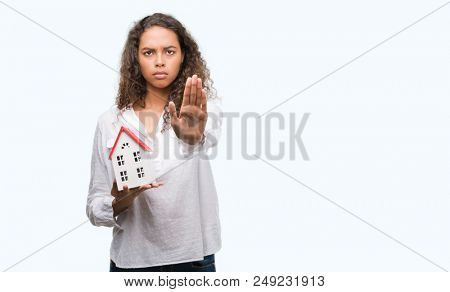 Young hispanic real state agent woman holding small house with open hand doing stop sign with serious and confident expression, defense gesture