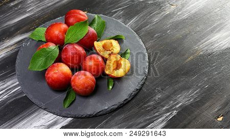 Red Plum Fruits On Branch With Green Leaves On A Round Shale Board A Dark Background. Top View, Spac