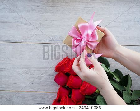 Preparing A Gift, On A Wooden Table, With Roses. Female Hand Holding A Gift, Wrapped With Ribbon, On