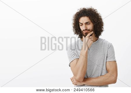 Studio Shot Of Doubtful Focused Adult Bearded Man With Dark Skin And Afro Hairstyle, Holding Hand On