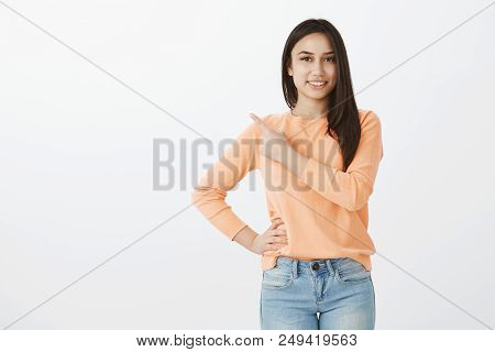 Portrait Of Attractve Tanned Female Coworker In Urban Outfit, Holding Hand On Hip And Pointing At Up