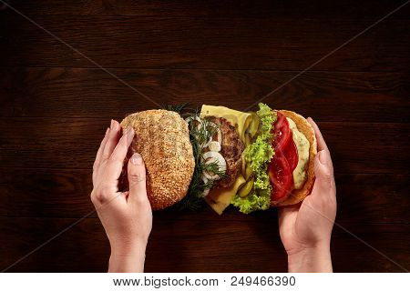 Woman's Hands Holding Traditional American Burger On Wooden Background, Top View, Close-up.