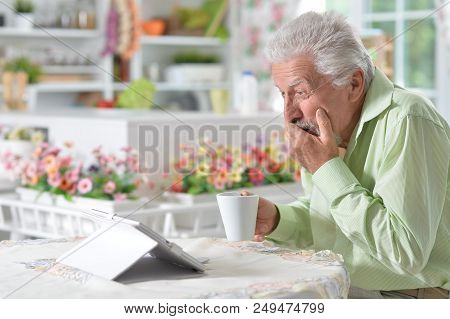 Emotional Senior Man Looking At Digital Tablet While Drinking Tea