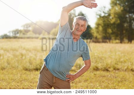 Outdoor Shot Of Active Senior Male Pensioner Bends Body, Does Physical Exercises In Coutryside, Prac
