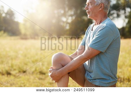 Cropped Shot Of Serious Mature Male Stretches Legs On Green Field, Looks Into Distance, Does Physica