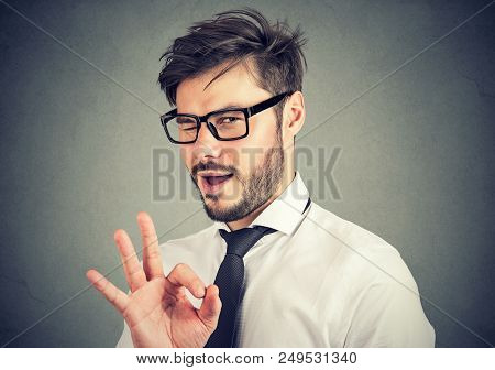 Young Bearded Man In Glasses Showing Ok Gesture And Blinking At Camera With Overconfidence
