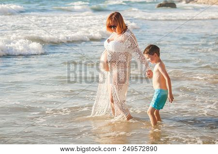 Beautiful Young And Pregnant Caucasian Woman Walking With Her Son On A Seaside Holding Him By His Ha