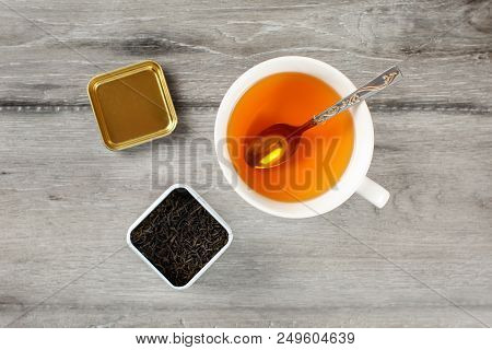 Table Top View On Cup Of Hot Amber Tea With Spoon, And Caddy Full Of Black Dried Loose Leaves, Place