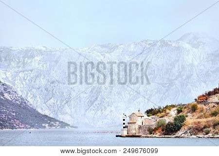 A Small Black And White Striped Lighthouse Near The Old Stone Chapel On The Background Of The Water 