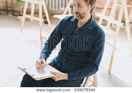 Warm Toned  High Angle Portrait Of Pensive Mature Man Sitting Alone In Loft Space And Drawing Sketch