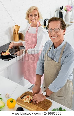High Angle View Of Senior Couple Cooking Together At Kitchen And Looking At Camera