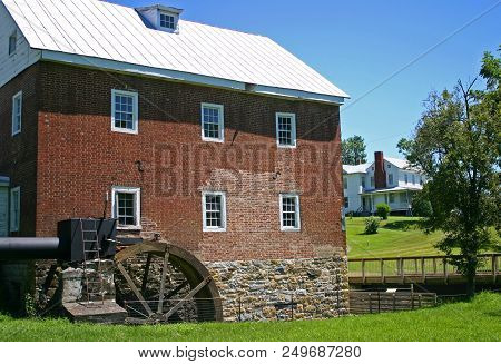 An Old, Brick, Two-story Gristmill Next To A Footbridge And A Farmhouse