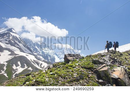 Mountain Landscape With Two Hikers On Peak Of High Hill Look At Snowy Mountains On Clear Sunny Summe