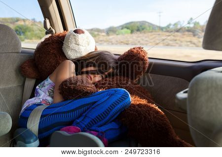 A Tired Little Girl Rides, Curled Up In Her Booster Seat Atop Of Her Big, Stuffed Bear, In A Minivan