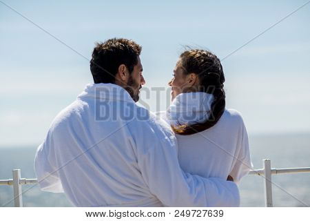 Back view of man in bathrobe embracing his wife during talk by waterside at summer resort