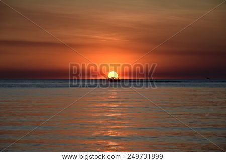 A Cruising Sail Boat Passes Majestically Beneath The Enchanting Sunset. The Sun Casts Orange Shades 