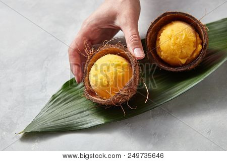 Two coconut shells with balls of apetitic mango ice cream on a green leaf on a gray concrete background with copy space for text. Girl's hand takes ice cream. Top view