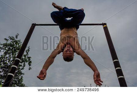 A Man With A Bare Torso In Blue Pants Upside Down Doing His Morning Exercises On A Horizontal Bar Ag